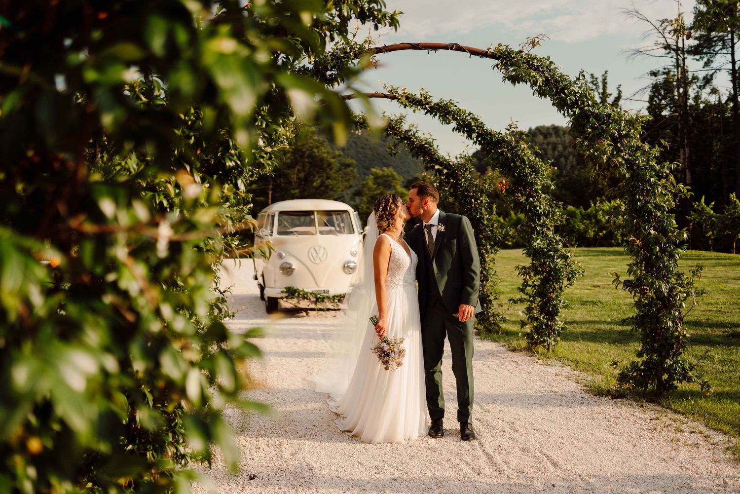 Bride and groom share a romantic moment under a lush floral arch with a vintage white VW van in the background.