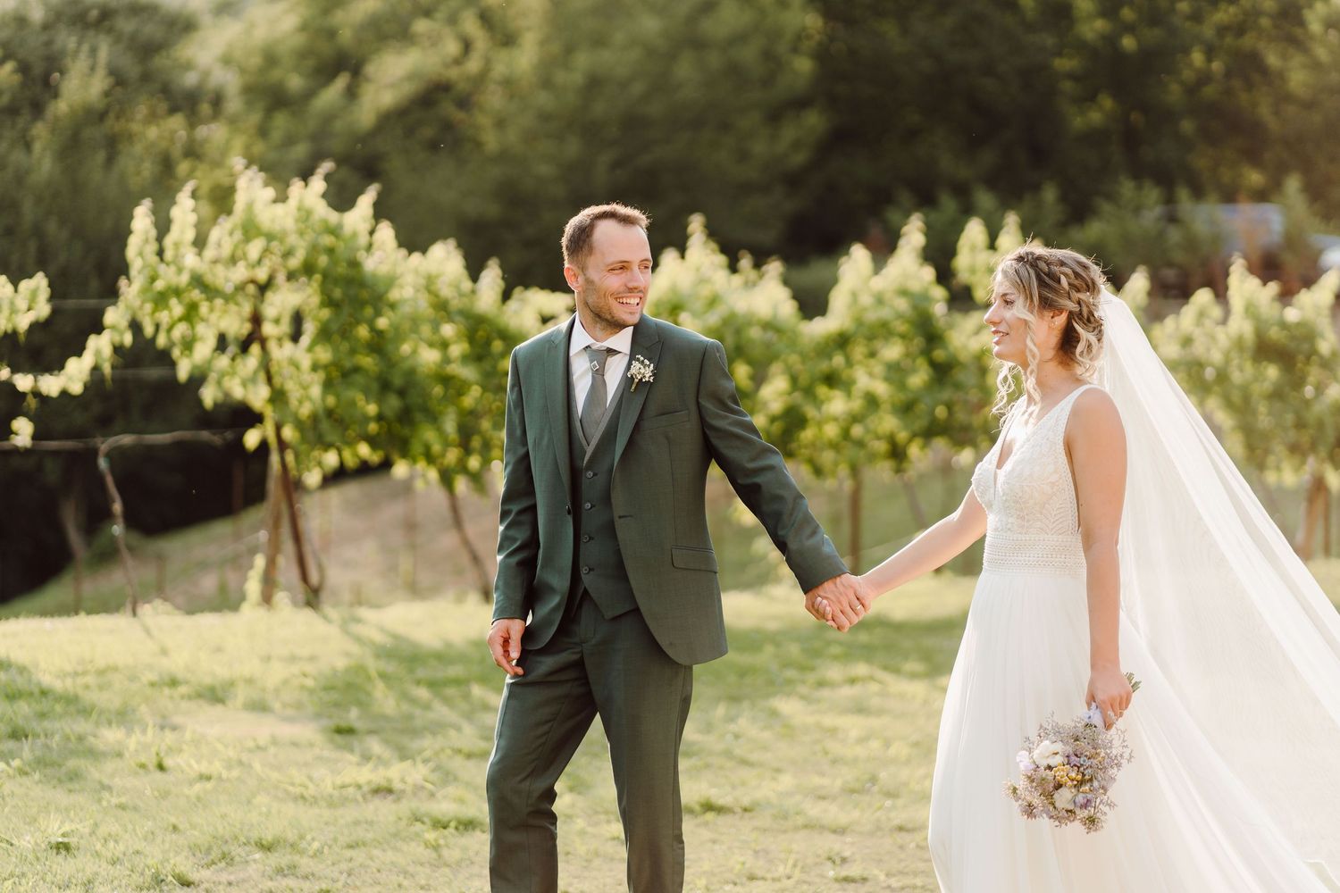 Bride and groom holding hands smiling at each other in a vineyard, groom in green suit, bride in white gown with veil.