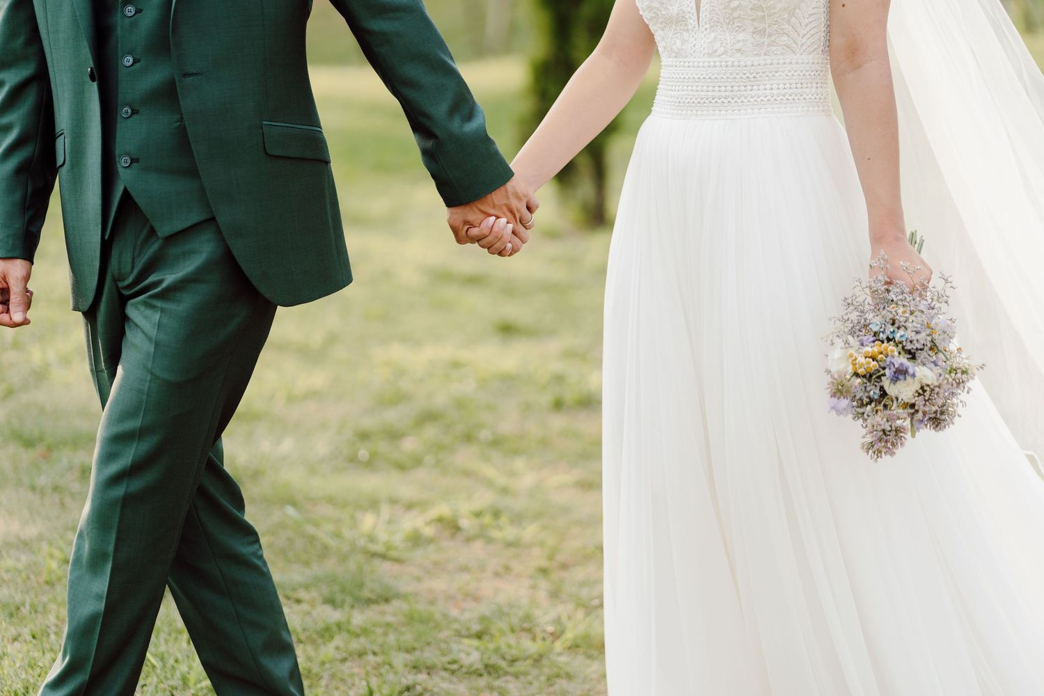 Bride in white gown holding wildflower bouquet walks hand in hand with groom in dark green suit outdoors.