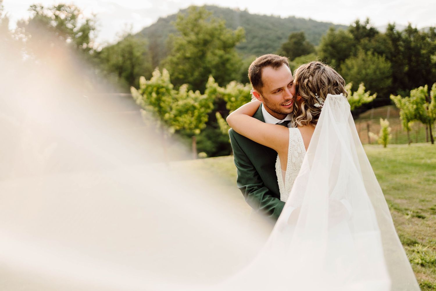 A bride and groom share a romantic embrace outdoors at a vineyard wedding, with her veil flowing in the breeze.