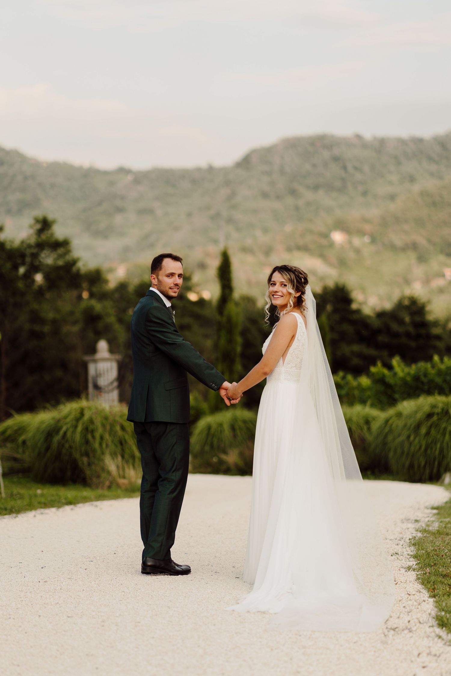 Bride and groom holding hands on a gravel path with lush green hills in the background on their wedding day.