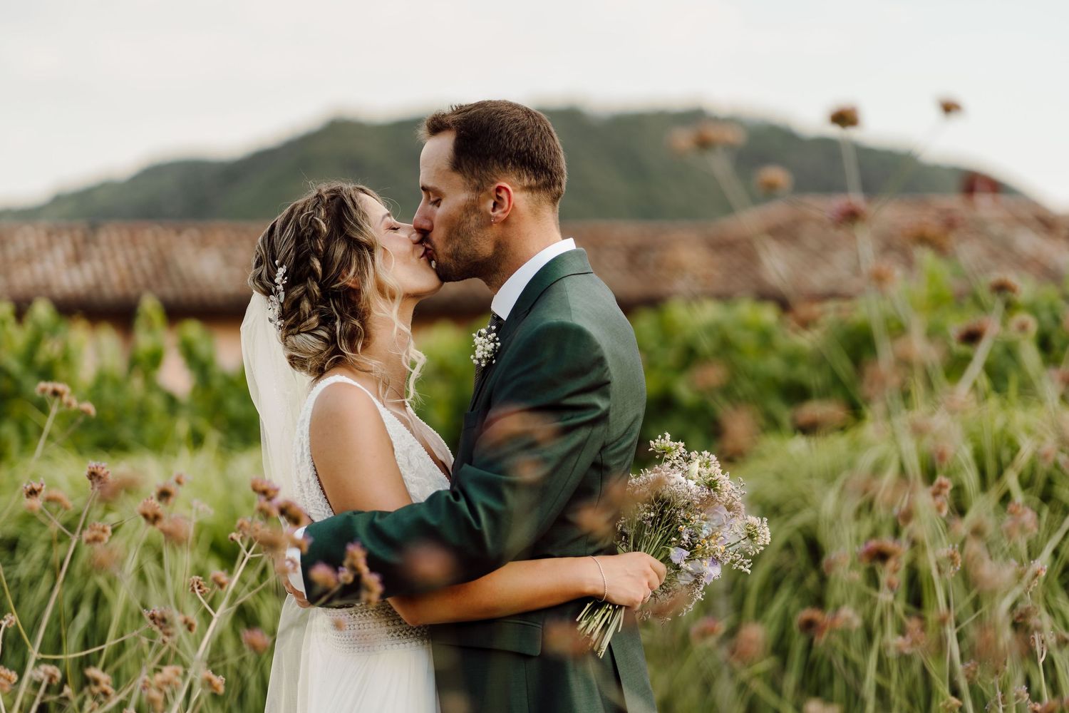 Bride and groom sharing a romantic kiss in a wildflower field with a scenic mountain backdrop on their wedding day.