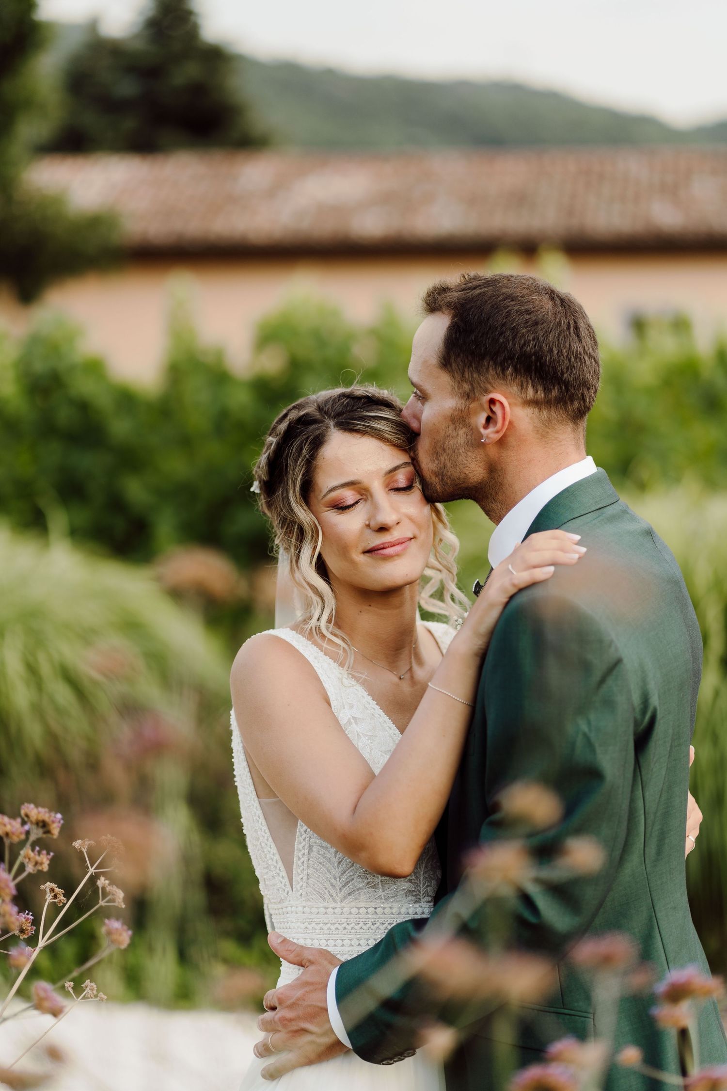 A groom in a green suit tenderly kisses his bride on the forehead in a lush garden setting.