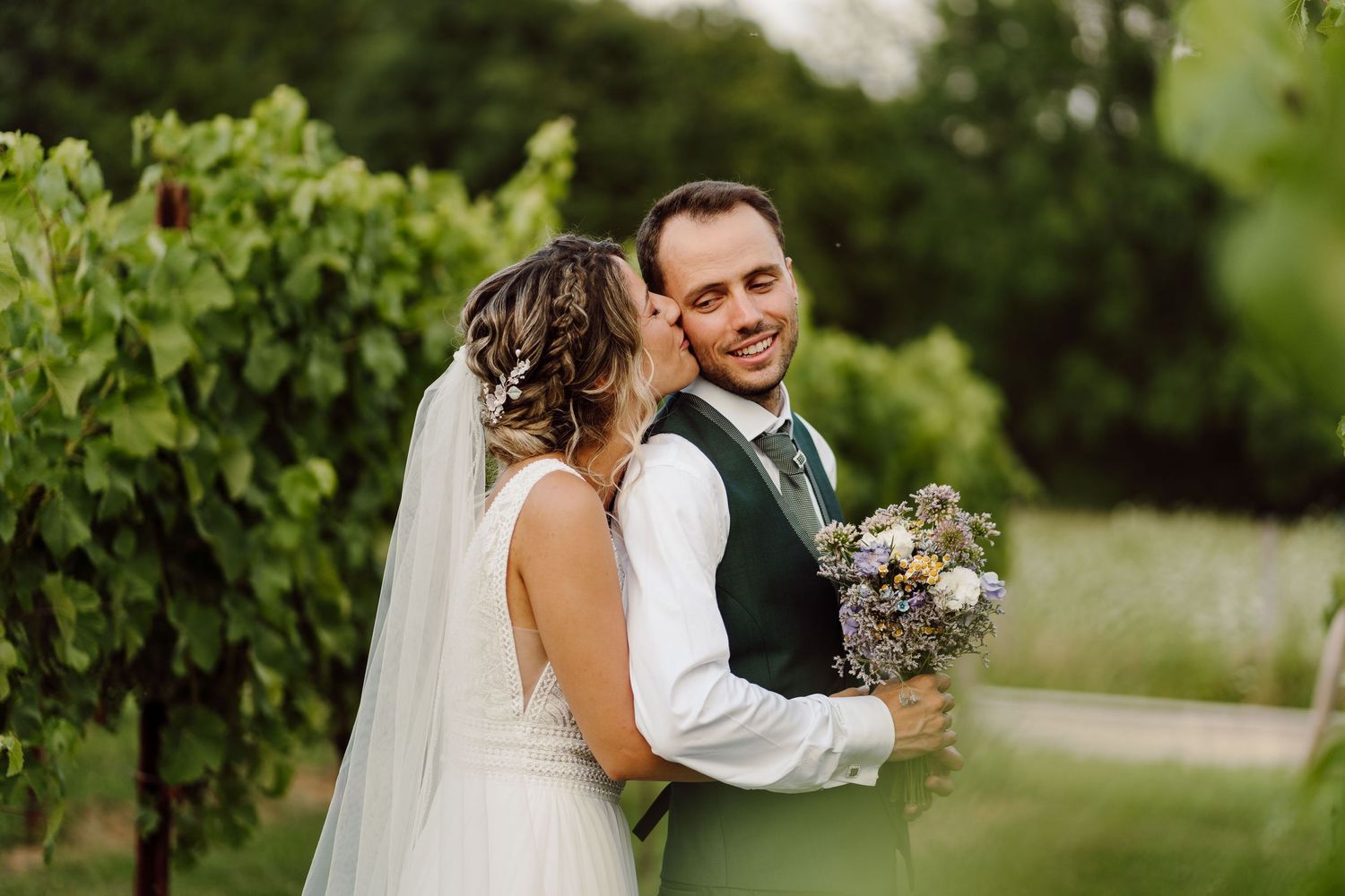 A bride and groom share a romantic moment in a lush vineyard, with the bride holding a bouquet of wildflowers.