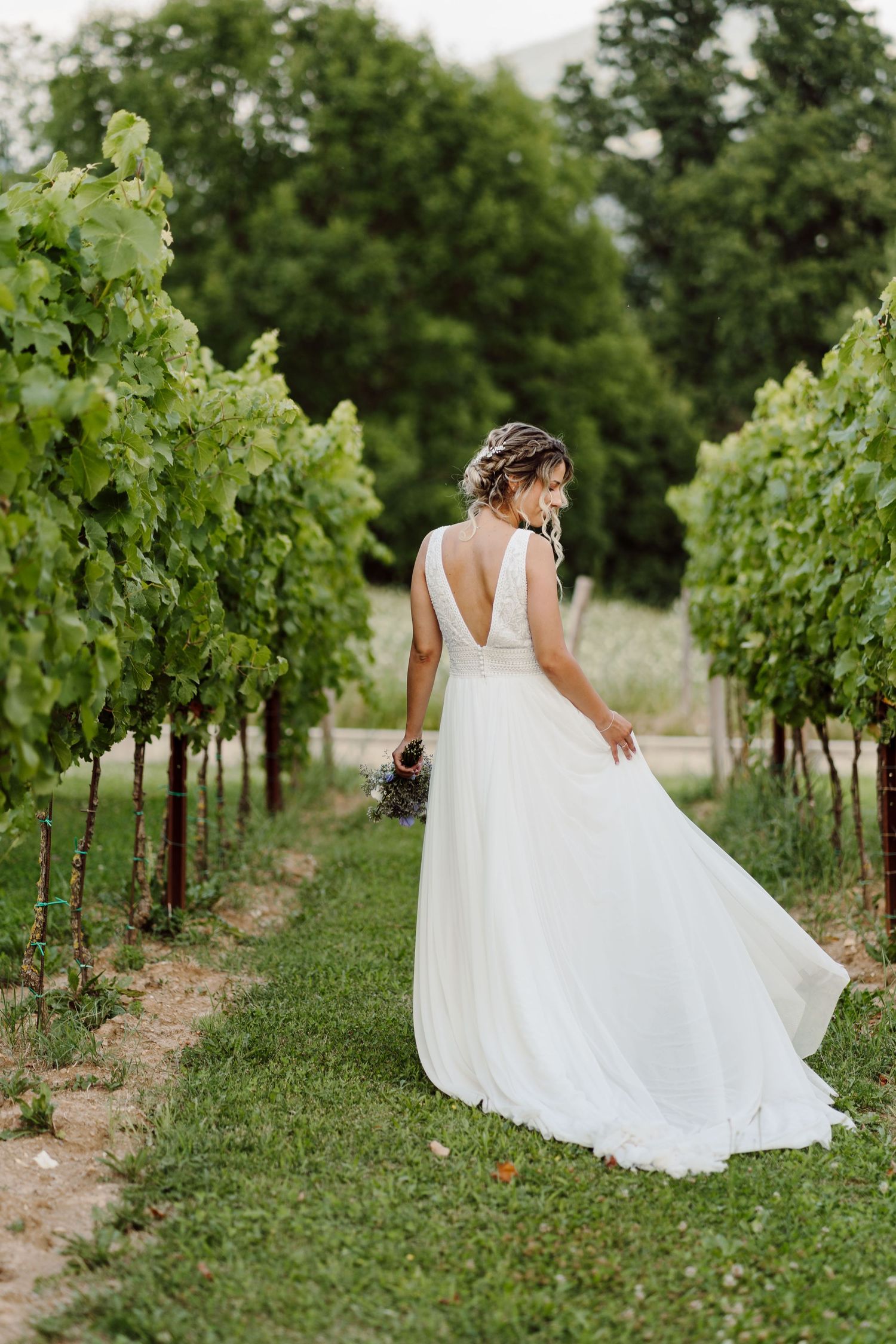 Bride in white gown walks through lush vineyard rows, holding bouquet, dress flowing gracefully in outdoor wedding setting.