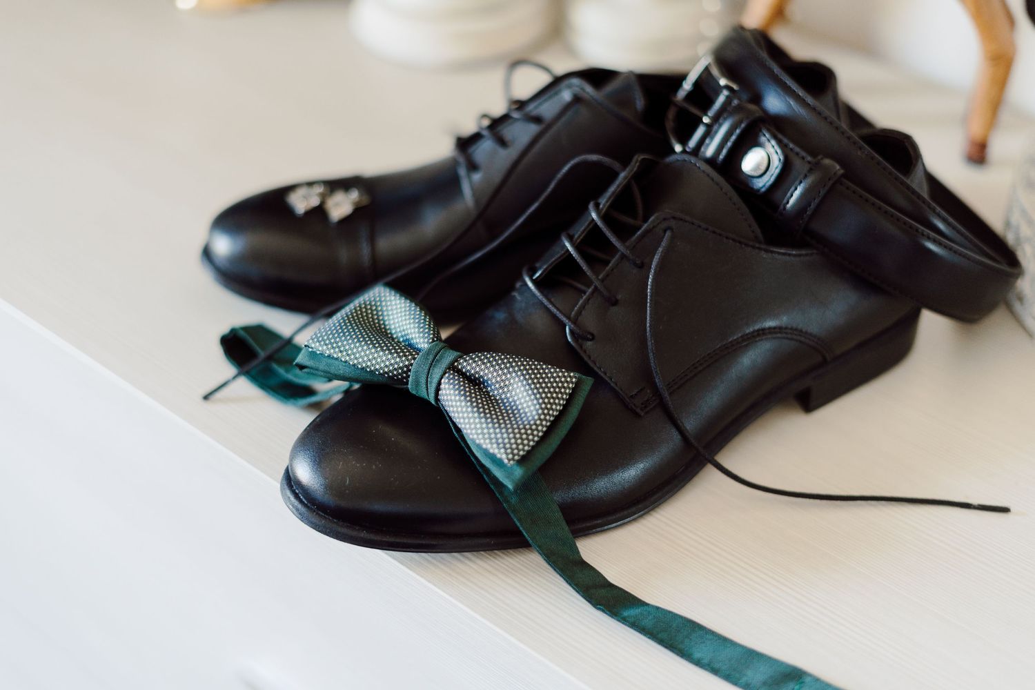 Black leather dress shoes with a green bow tie resting on a white surface, perfect for a formal wedding.
