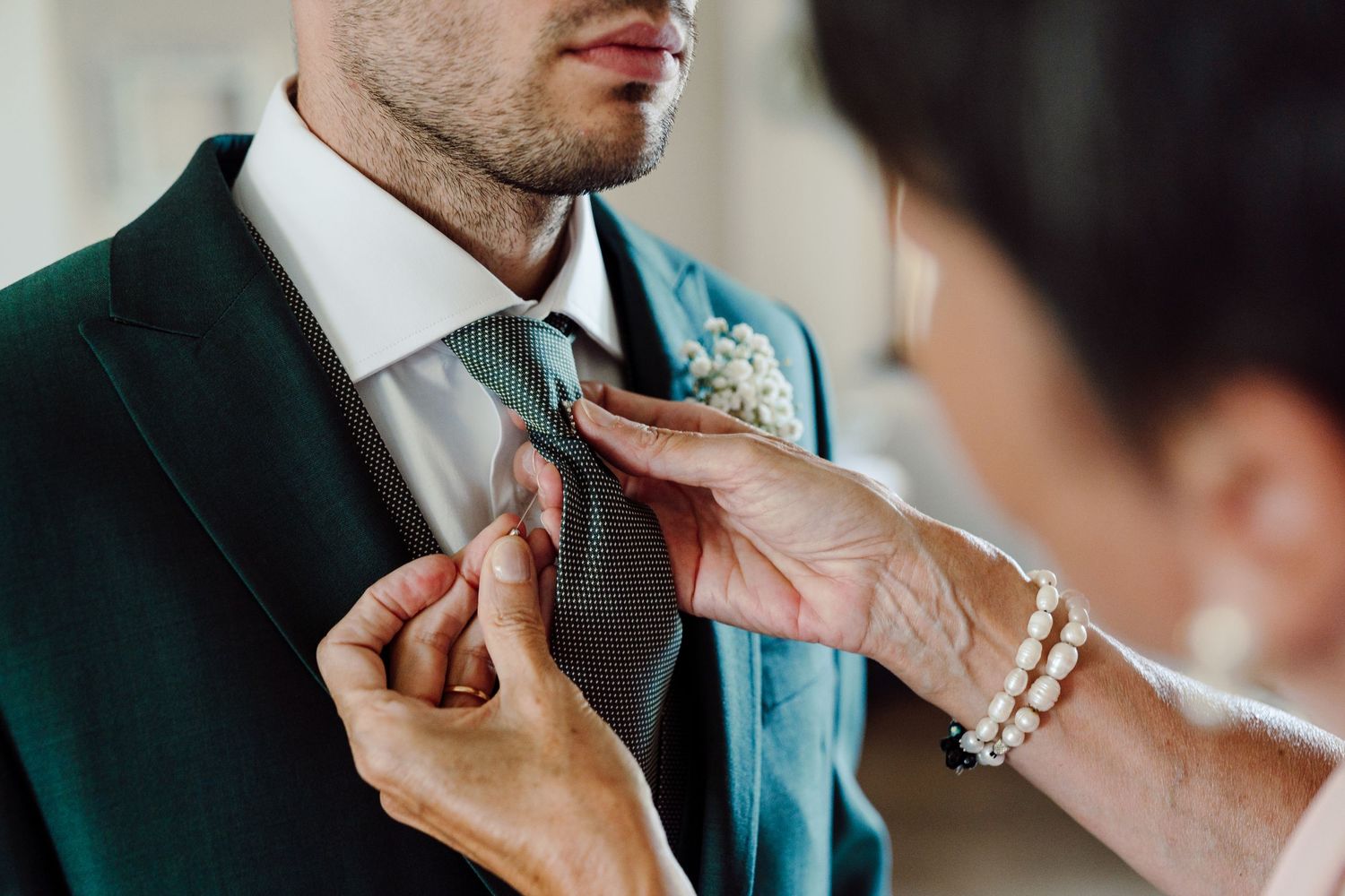Woman adjusting groom's green tie and boutonniere on wedding day suit preparation.