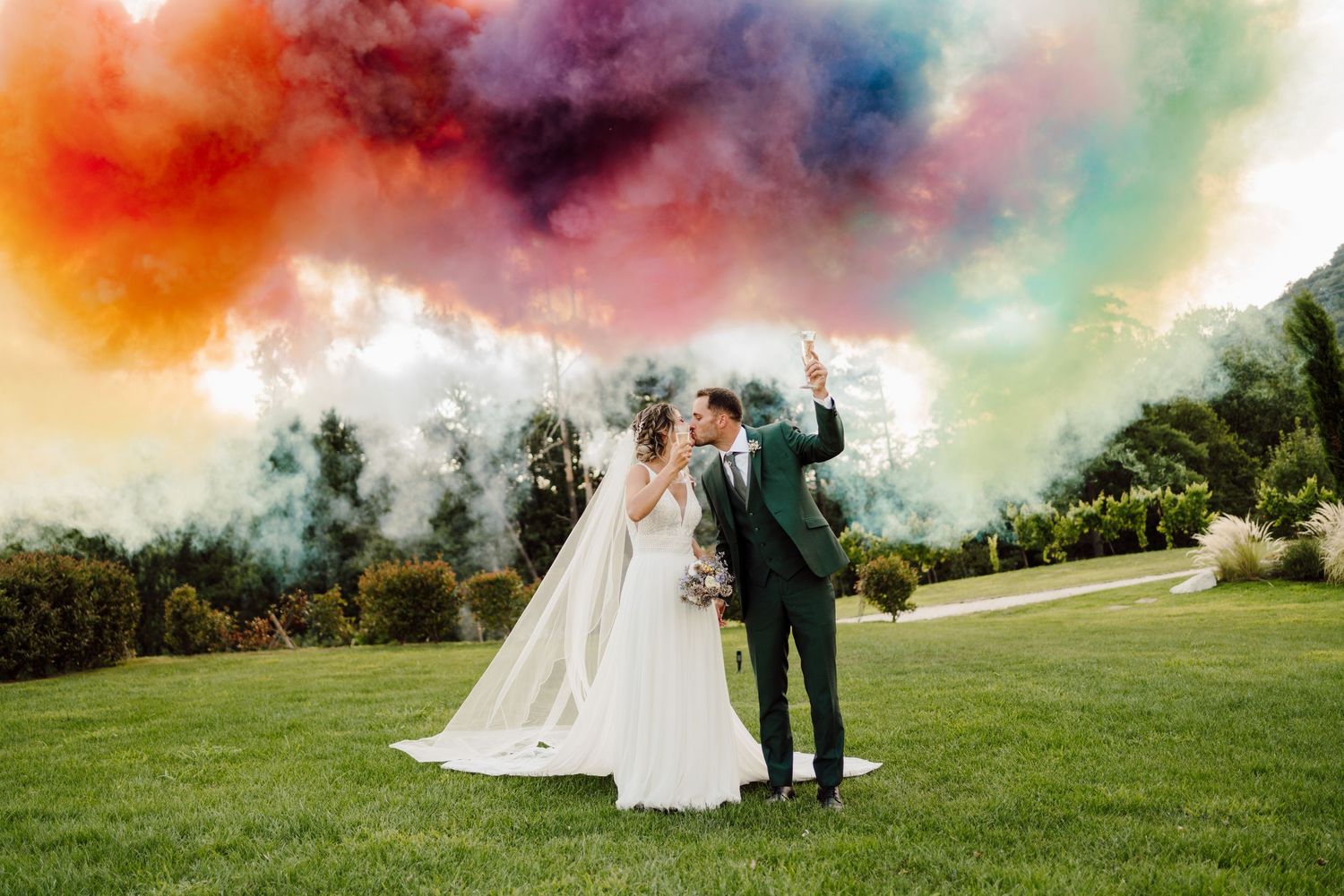 Bride and groom kissing on green lawn under vibrant rainbow colored smoke clouds at outdoor wedding celebration.