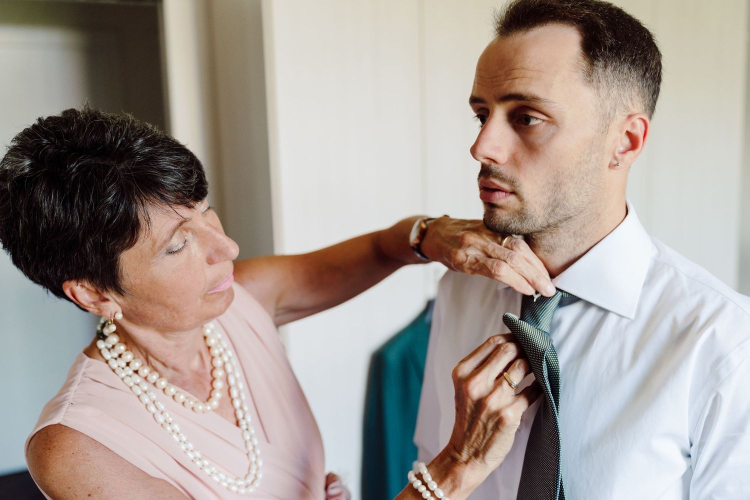 Older woman adjusting groom's tie before wedding ceremony, wearing pearl necklace and pink dress.