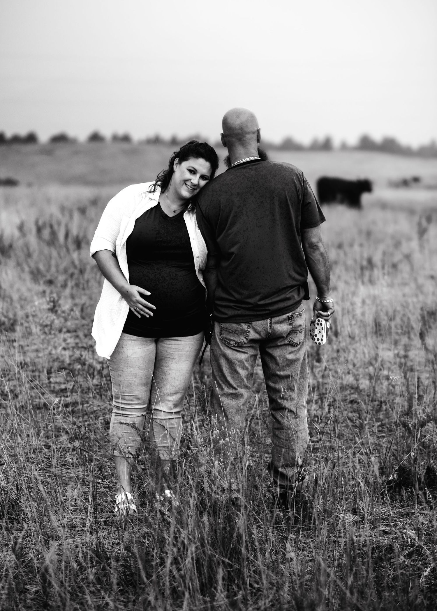 Black and white photo of a pregnant woman and bald man standing in a grassy field with cattle in the background.
