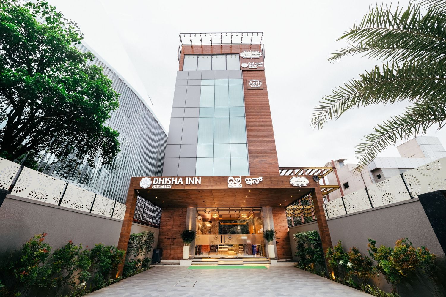 Modern Dhisha Inn hotel exterior with glass facade, brick accents, and palm trees at entrance.