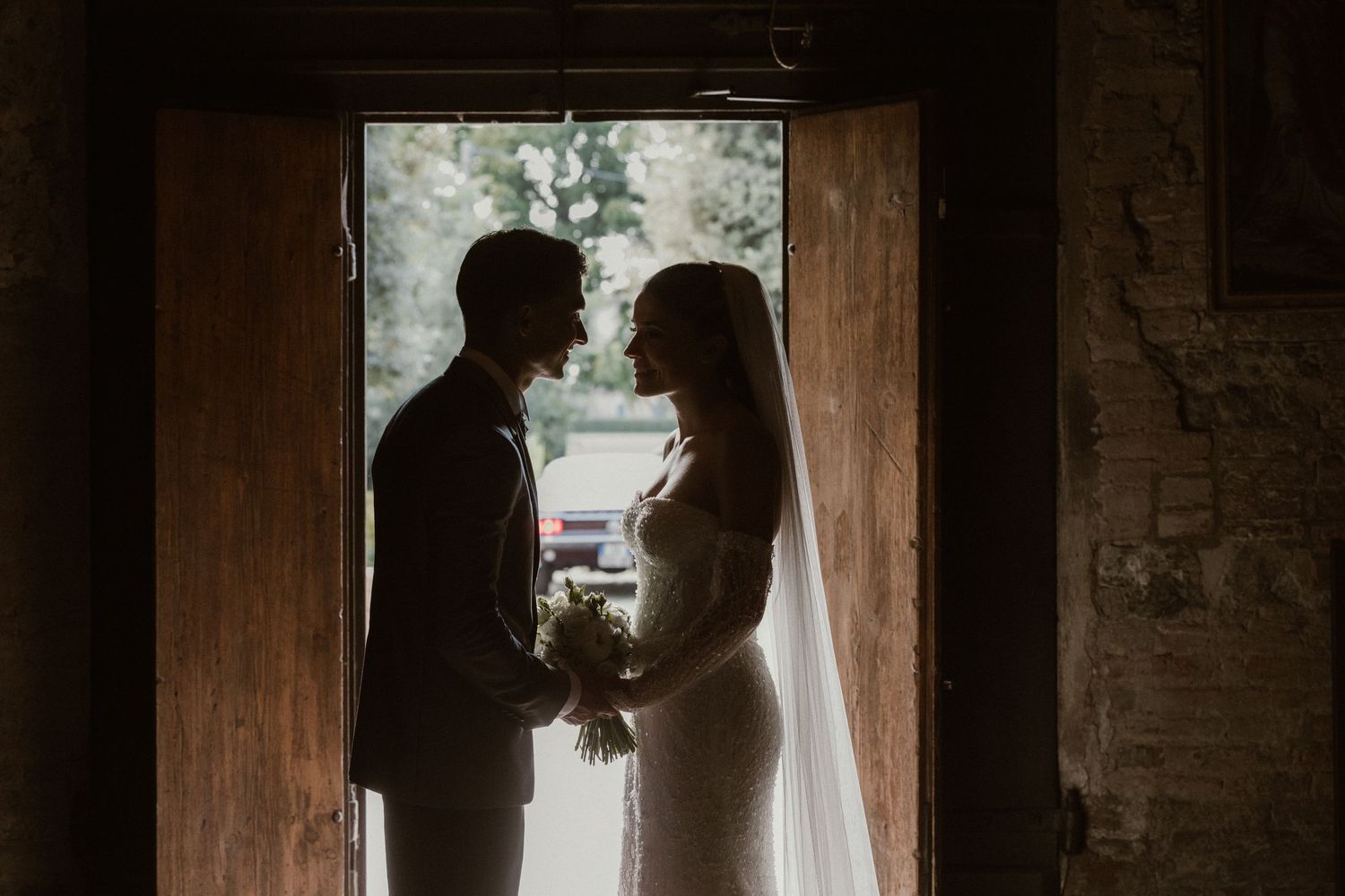 A bride and groom share a romantic kiss outdoors, surrounded by sunlit trees with beautiful light rays streaming through.