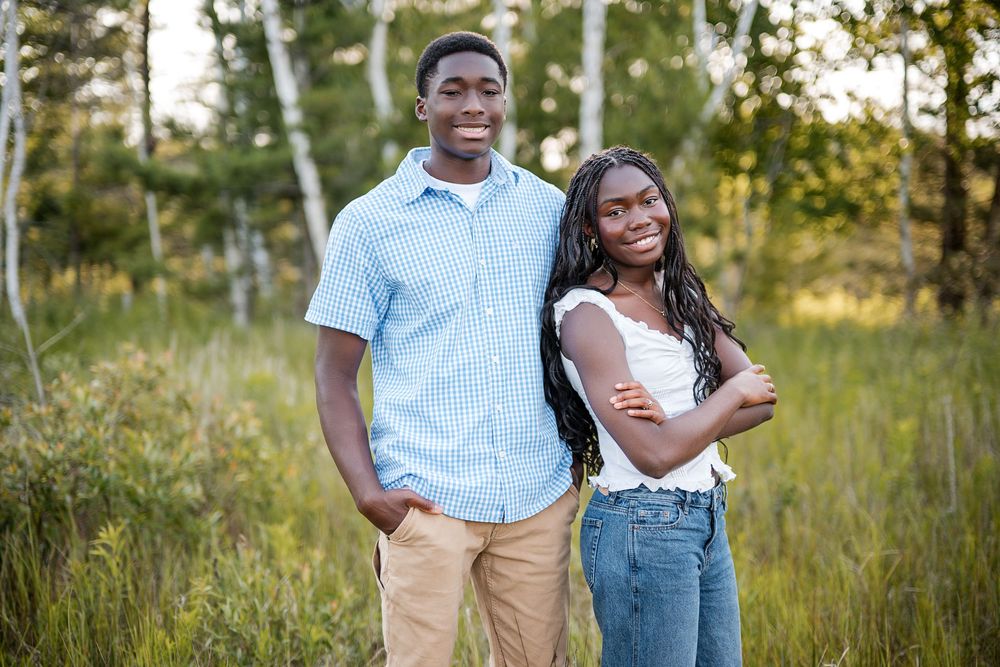 Two smiling young Black teens pose together in a sunlit field with green trees in the background.