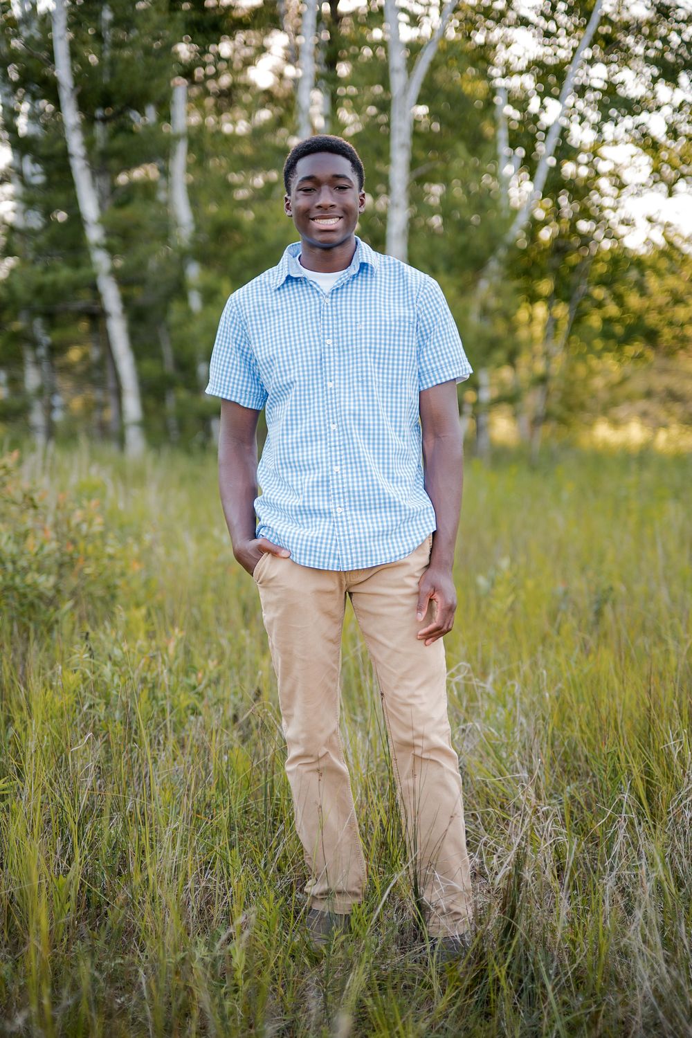 Young Black man smiling in a light blue plaid shirt and khaki pants, standing in a lush green field with birch trees.