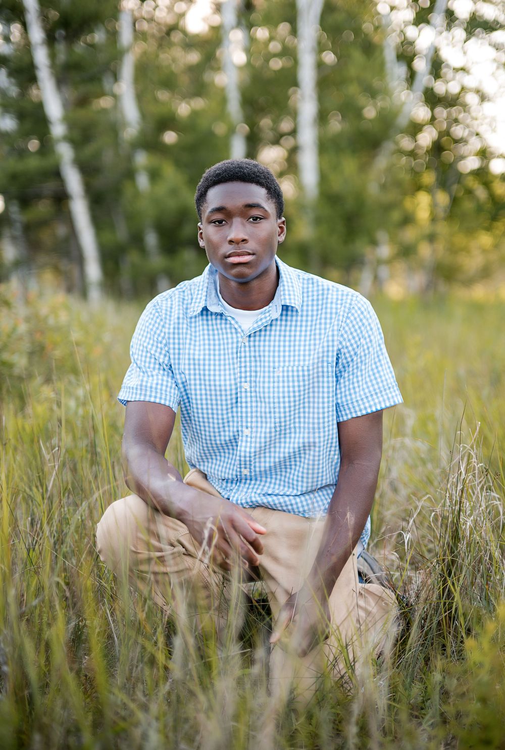Young man in blue checkered shirt and khaki pants sitting in tall grass with trees in background.