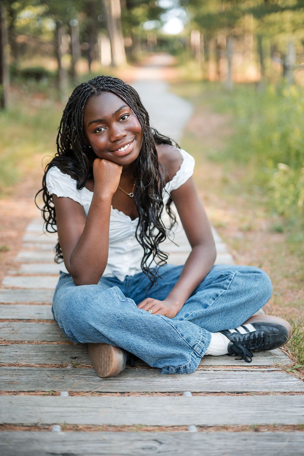 Smiling young Black woman with braids sits cross-legged on a wooden path in jeans and white top.