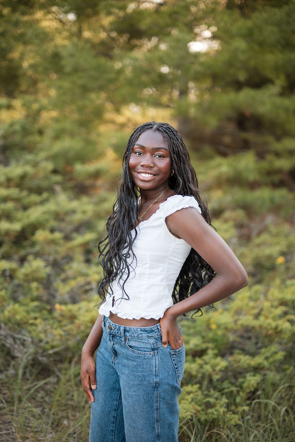 Smiling young Black woman with long braids wearing white crop top and jeans posing outdoors in a green wooded setting.