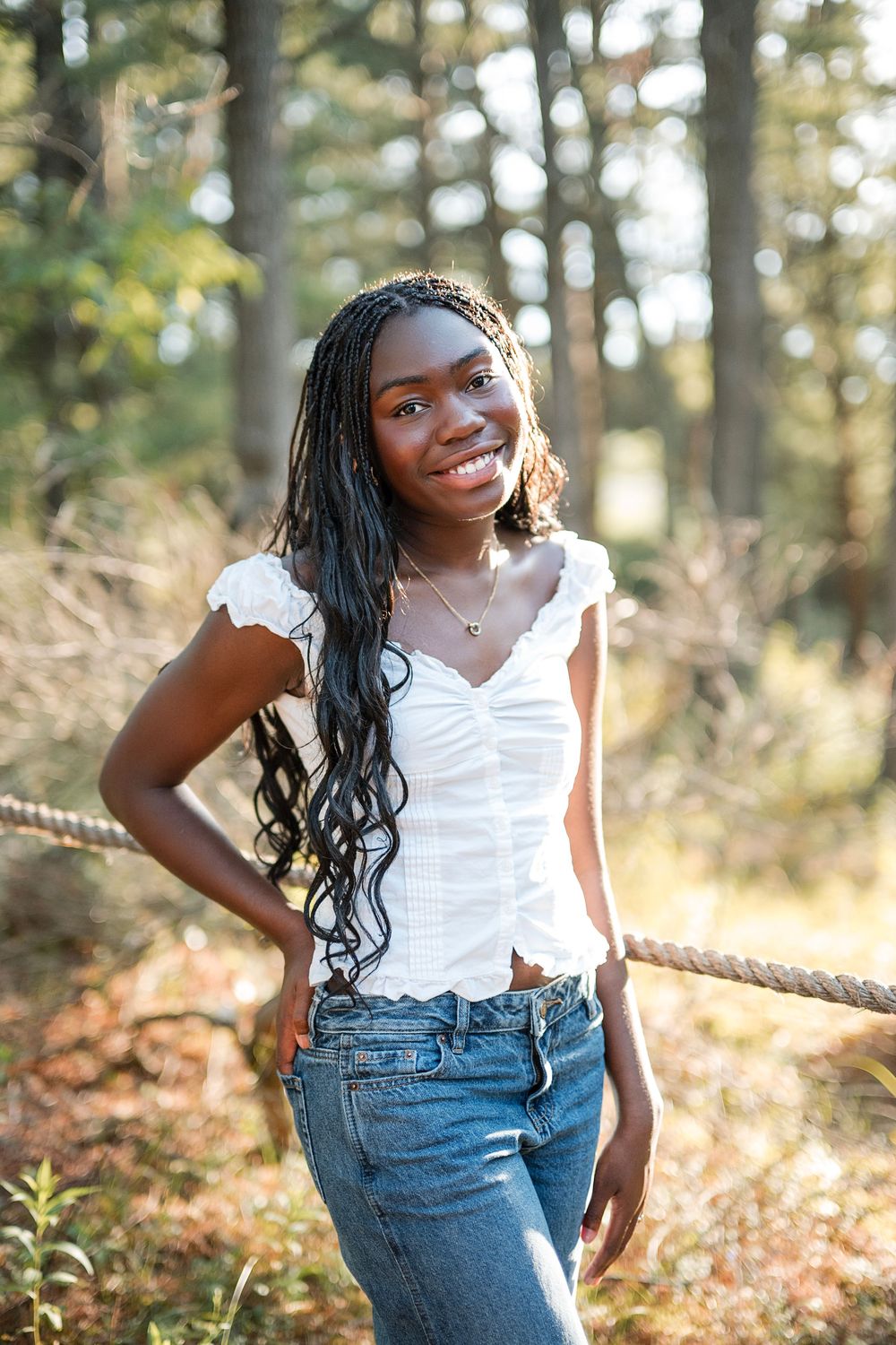 Young Black woman with long braids smiling in white top and jeans outdoors in a sunlit forest setting.