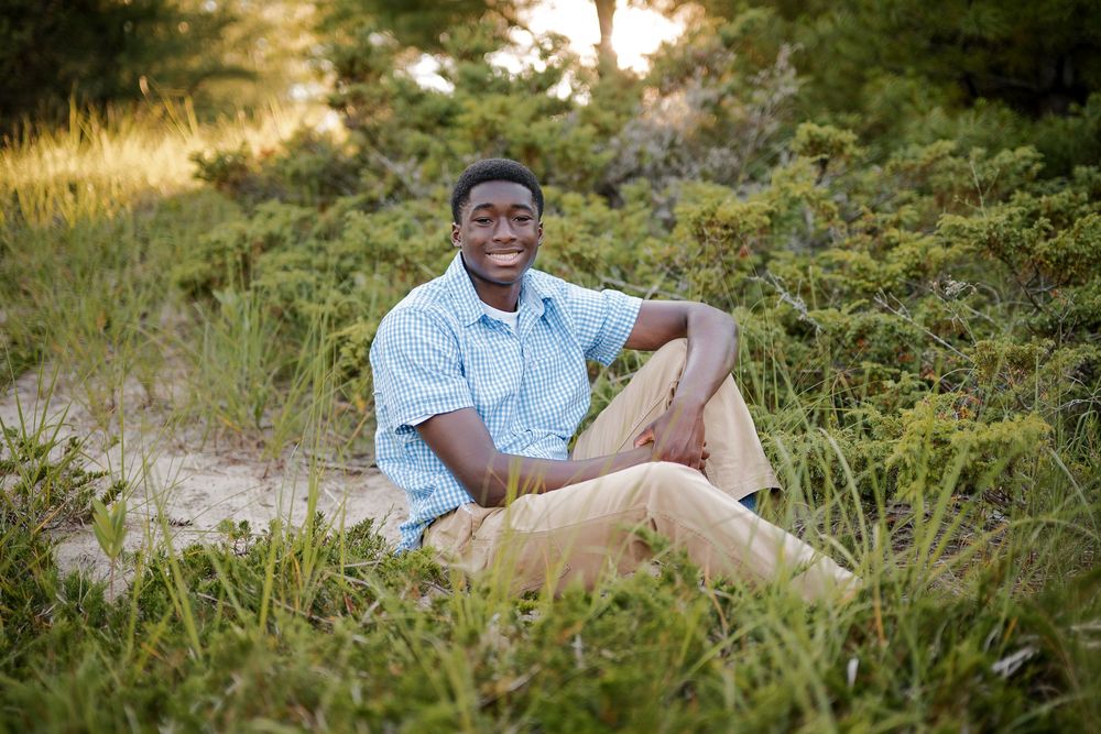 A young man smiles while sitting outdoors in lush green grass and sandy dunes during golden hour.
