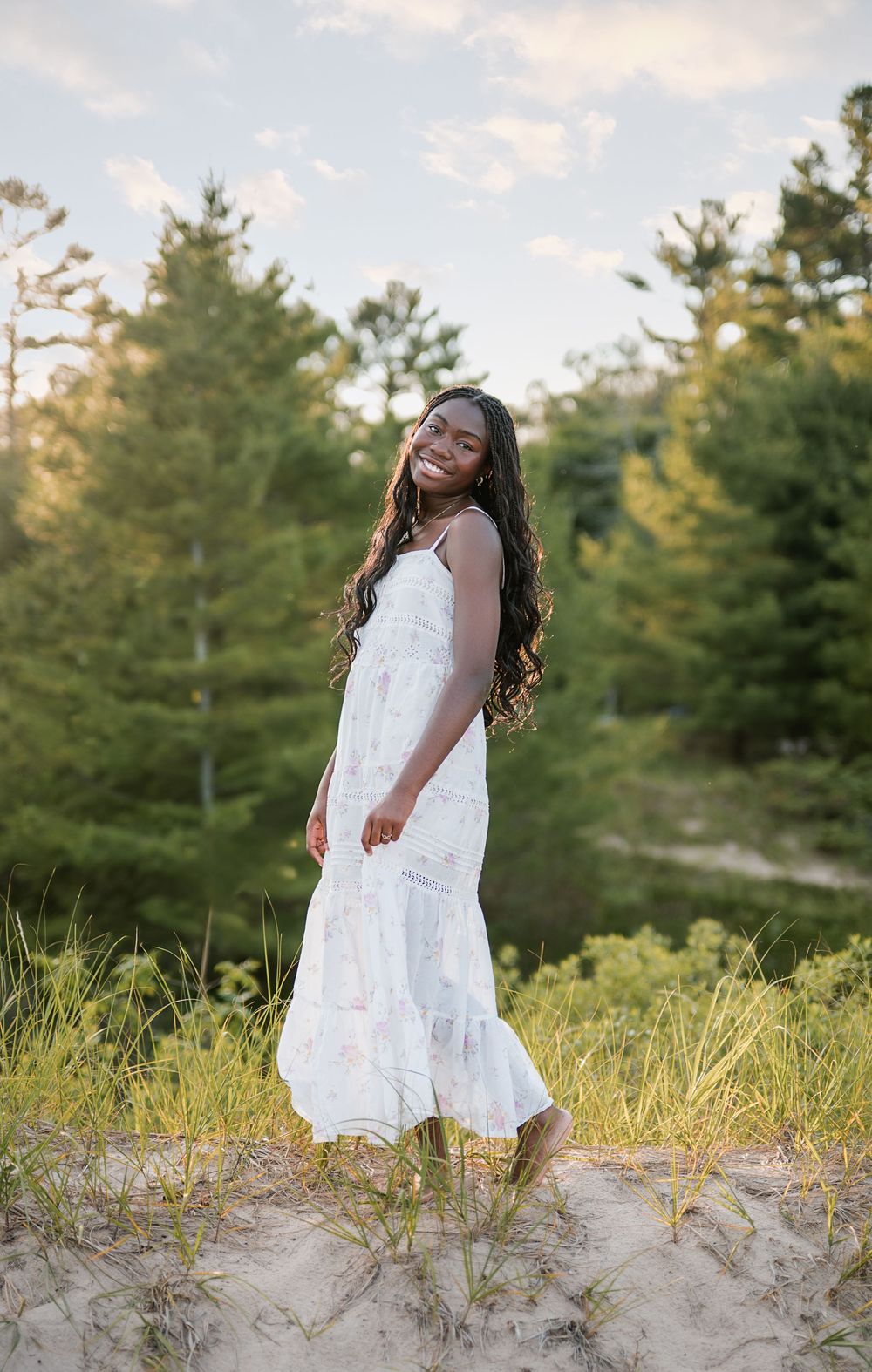 Young Black woman in white sundress smiling on sandy dunes surrounded by green trees at golden hour.