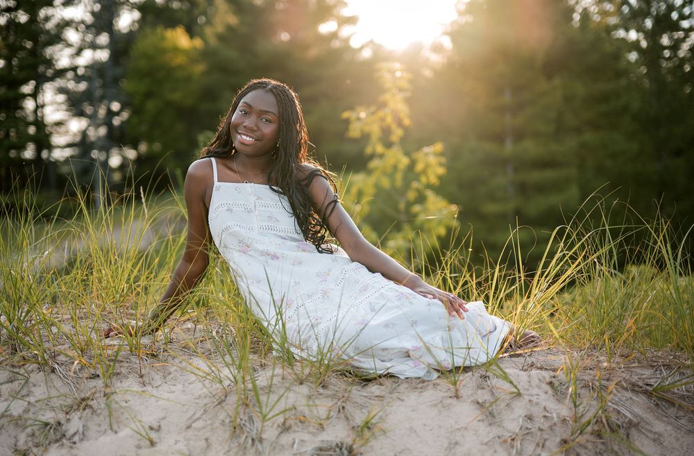 Young Black woman in white dress sitting on sandy dunes, smiling at sunset with lush green trees behind her.