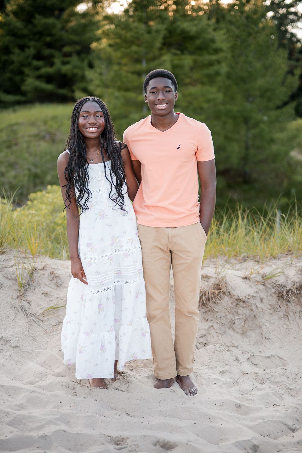 Two smiling young adults pose together on a sandy beach, wearing casual summer attire with green trees in the background.