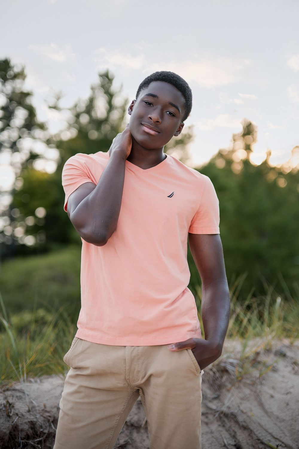 Young Black teen in a peach Nautica t-shirt and khaki pants poses outdoors with greenery in the background.