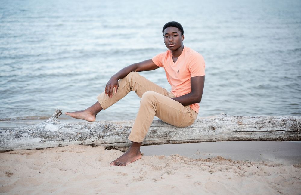 Young Black man in salmon polo and khaki pants sits barefoot on driftwood log at sandy beach with calm water behind him.