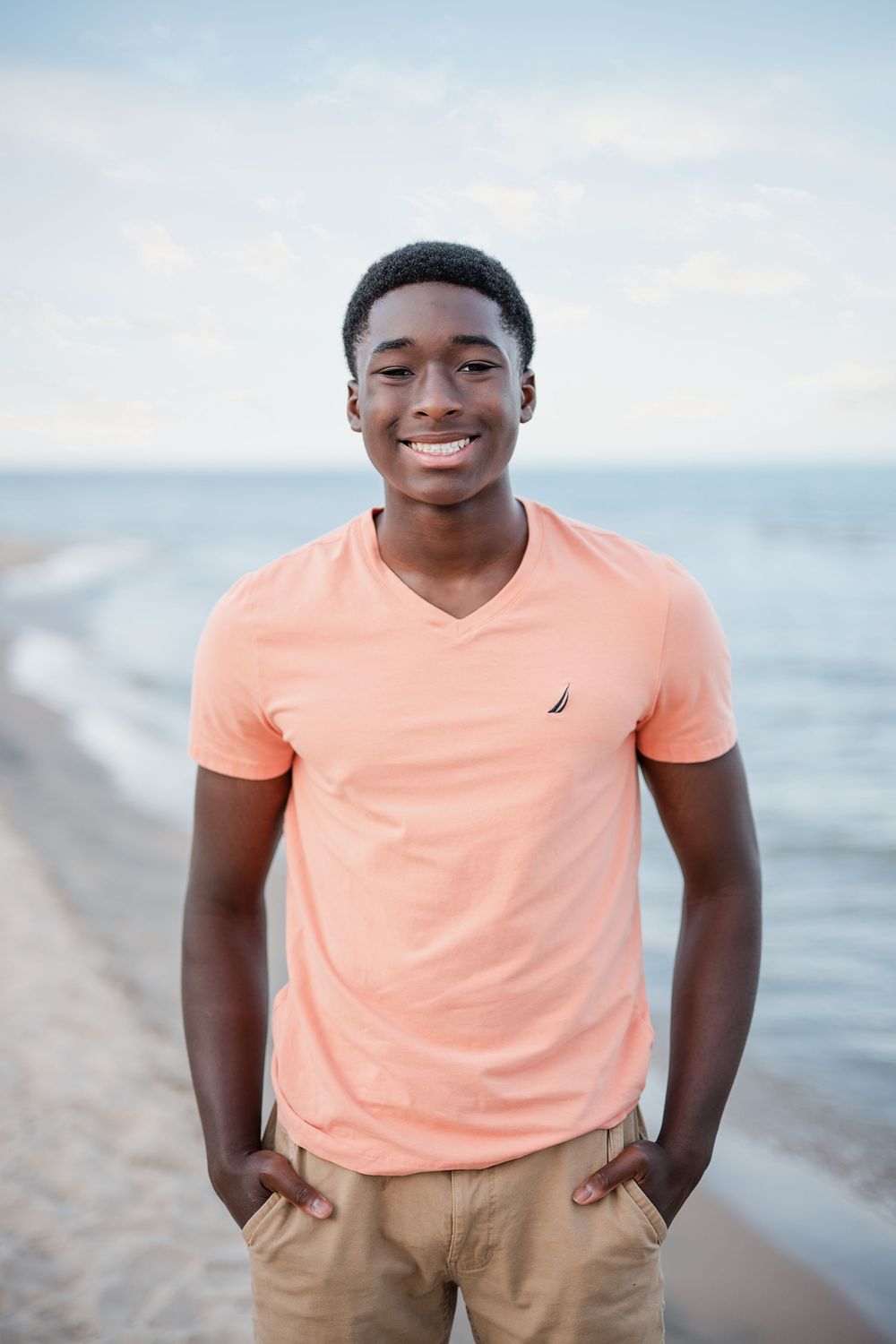 Smiling young Black man in a peach Nautica t-shirt and khaki pants standing on a beach.