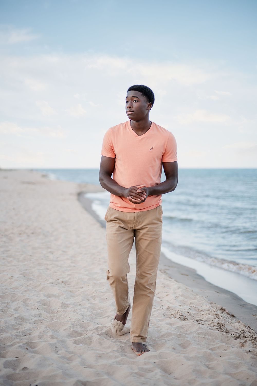 Young Black man in salmon t-shirt and khaki pants walking barefoot on sandy beach with ocean waves behind him.