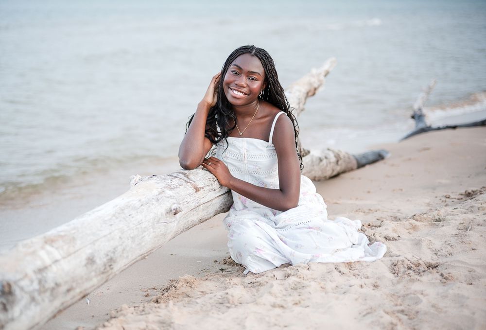Young woman in white dress smiling on sandy beach, leaning against driftwood log with ocean in background.