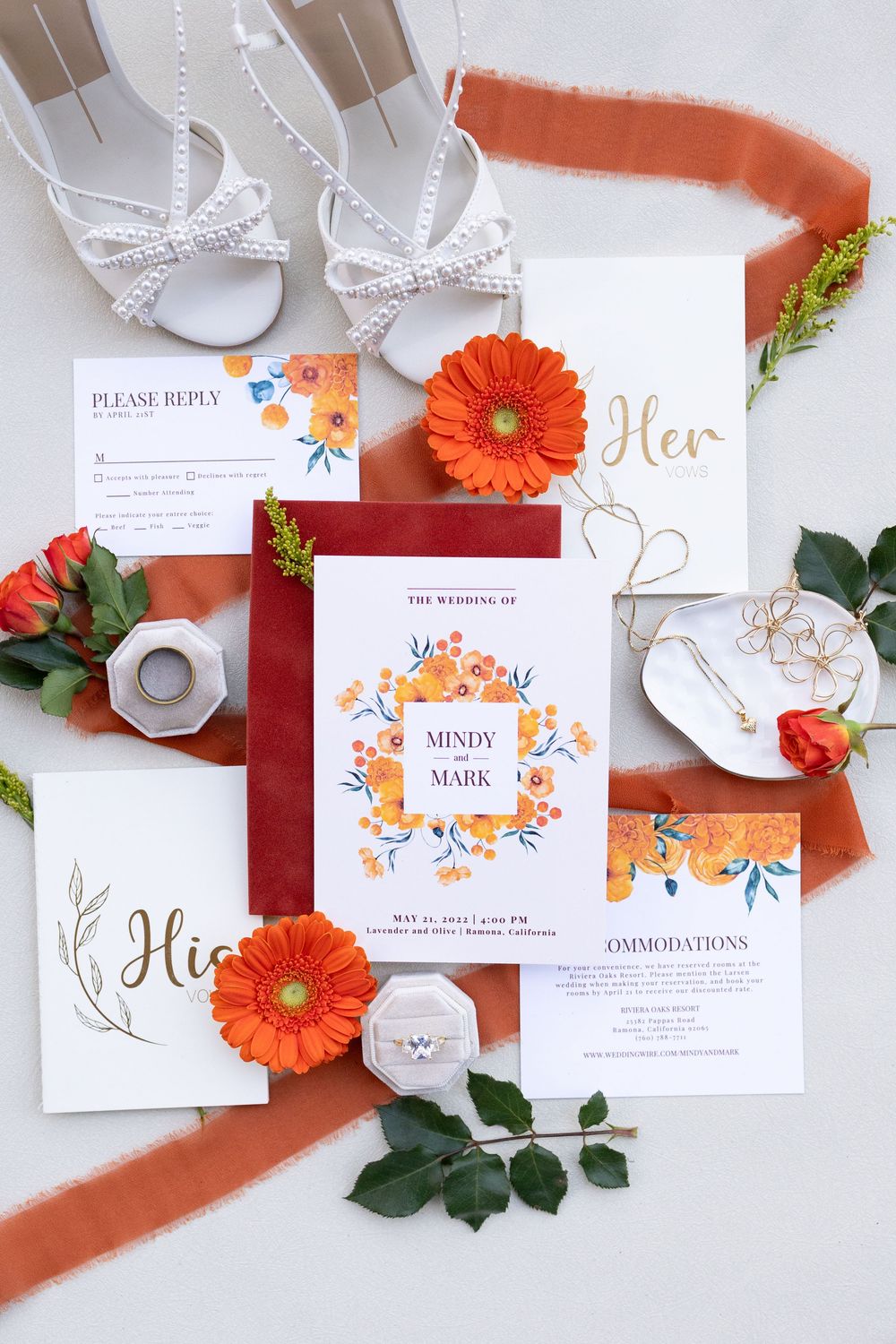 Elegant wedding invitation suite with orange floral design, white heels, gerbera daisies, and terracotta ribbon flatlay.