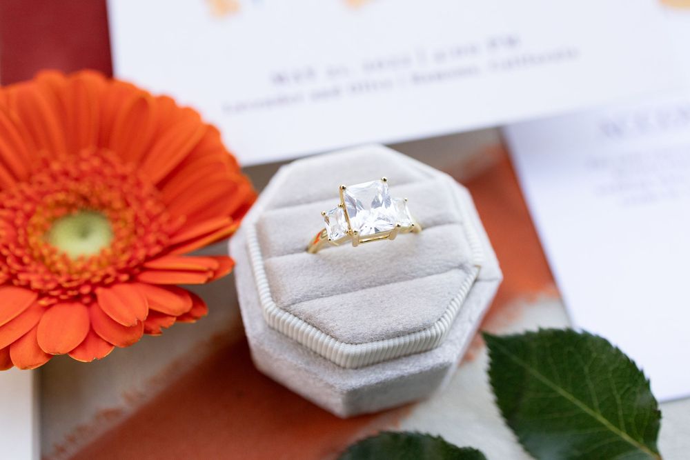 Gold three-stone emerald cut engagement ring displayed in a velvet ring box beside an orange gerbera daisy.