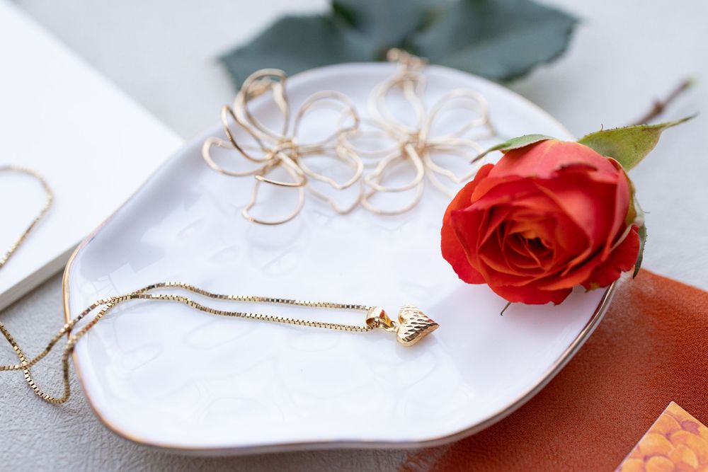 Gold pendant necklace with heart charm displayed on white ceramic tray beside a red rose and twine bow.