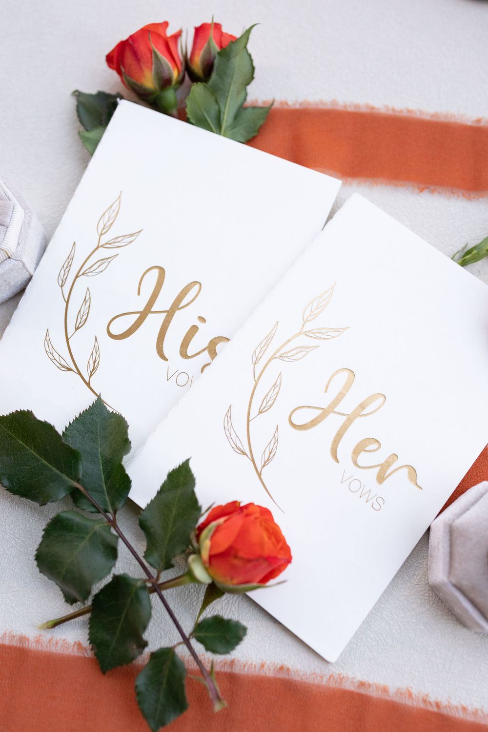 Gold foil His and Her vows cards with orange roses and ribbon on white background.