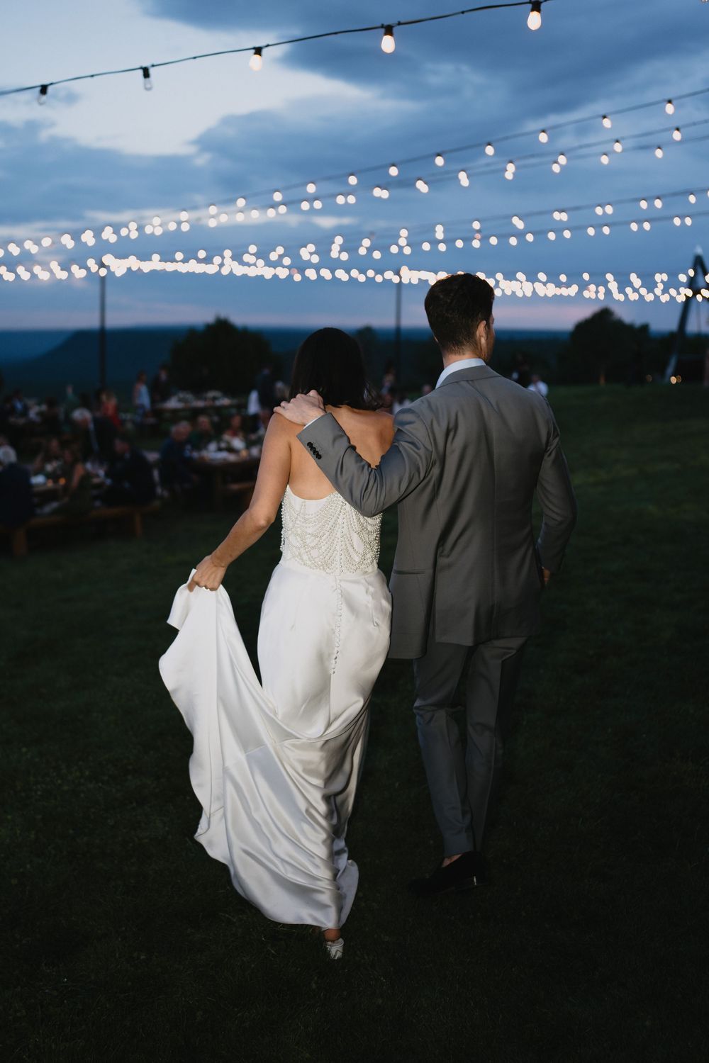 Bride and groom walking arm in arm at dusk under string lights at an outdoor wedding reception.