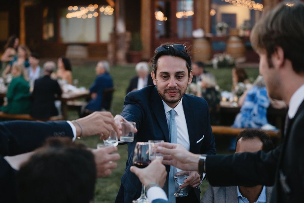 Man in navy suit toasting with guests at an outdoor wedding reception celebration.