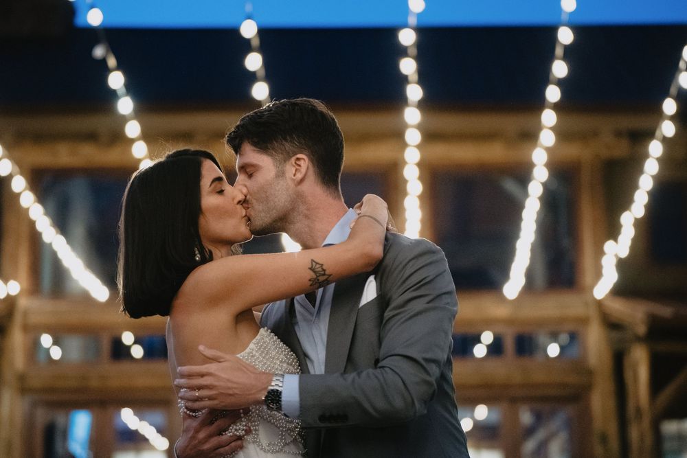 A couple shares a romantic kiss during their wedding reception under warm string lights in a rustic barn venue.