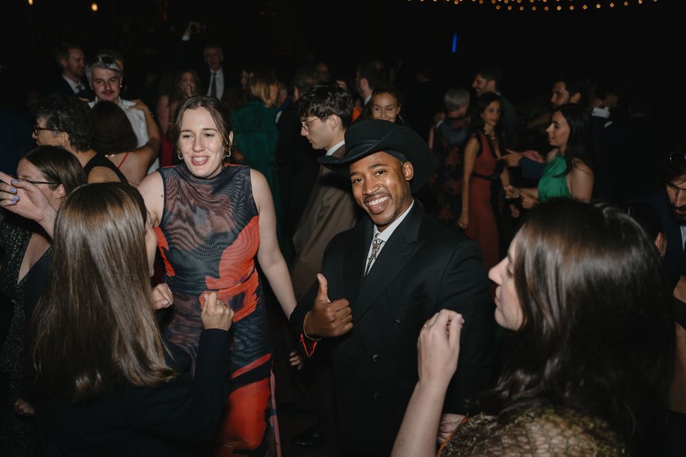 Guests dancing and smiling at a formal party event, man in black suit giving thumbs up on crowded dance floor.