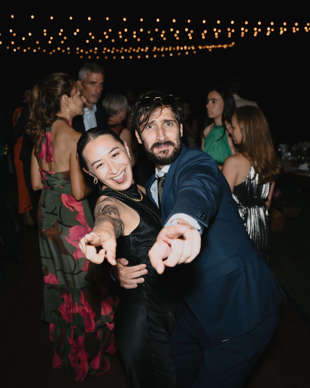 Two guests dancing and pointing at camera at an outdoor evening wedding reception with string lights.
