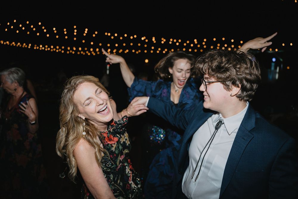 Three people dancing joyfully at an outdoor evening event with string lights in the background.