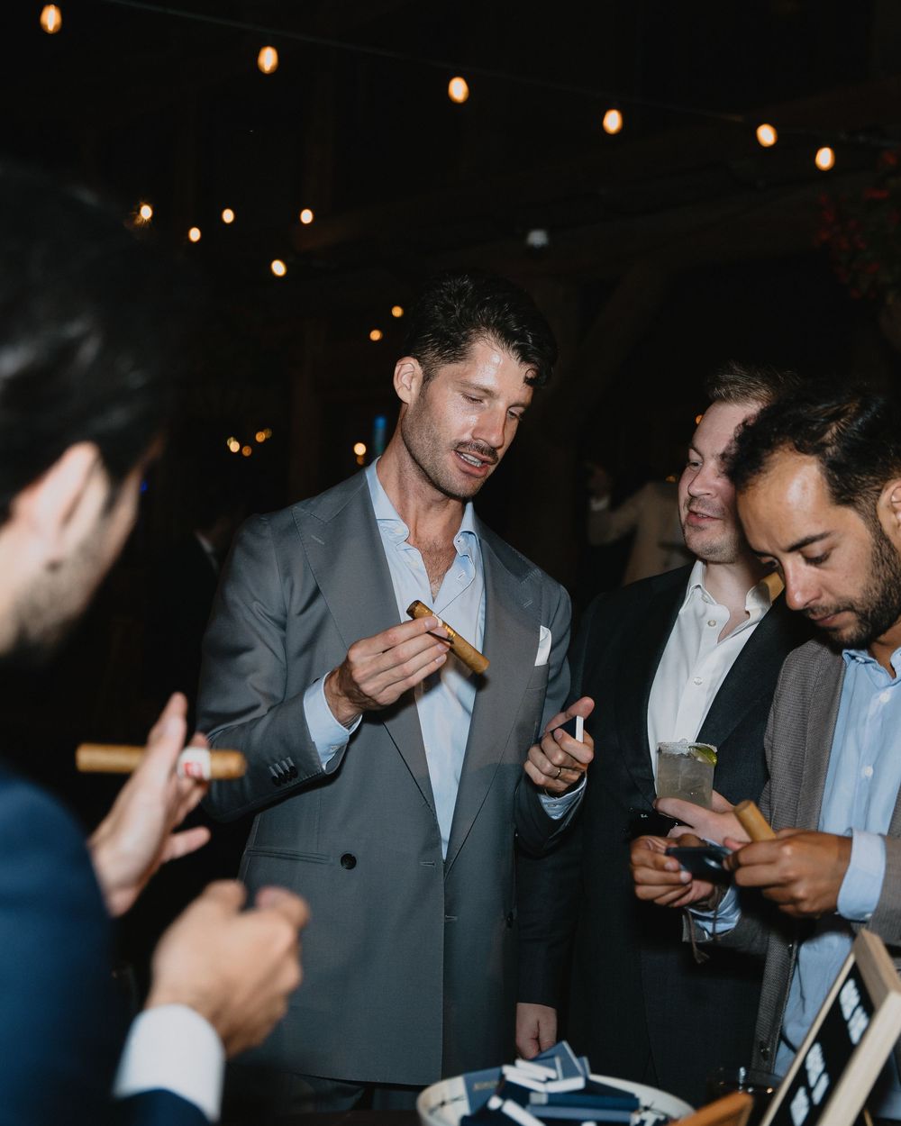 Man in gray suit holding a cigar at an outdoor evening party with string lights.