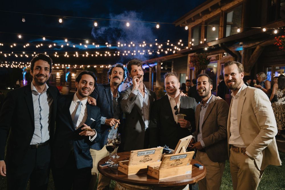 Group of well-dressed men smiling at an outdoor evening event with string lights and cigars on a wooden table.