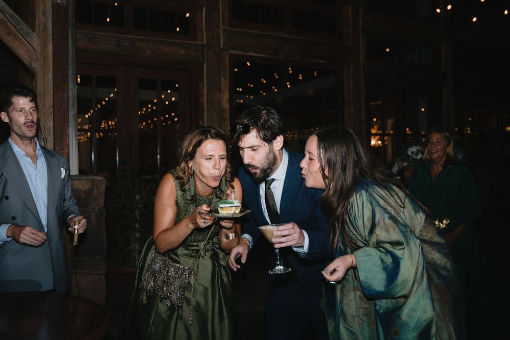 Guests in formal attire gather at a rustic barn wedding reception, sharing cake and drinks under warm string lights.