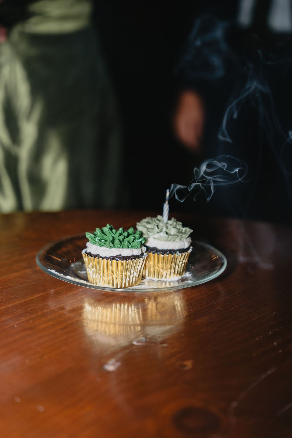Two decorated cupcakes with green frosting on a glass plate, one with a just-blown-out candle emitting smoke, on a wooden table.