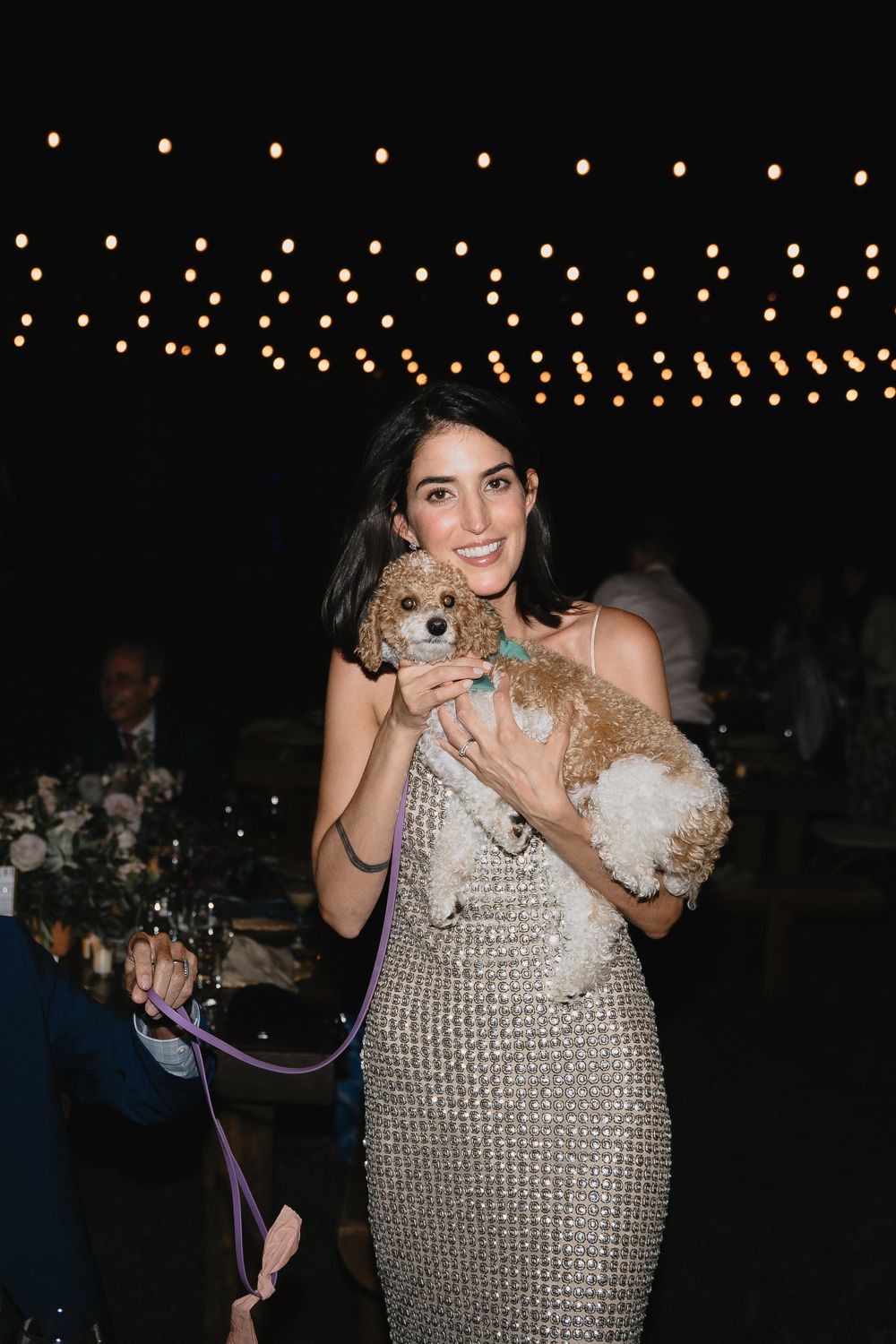 Woman in silver sequin dress holding a small dog at an outdoor evening event.