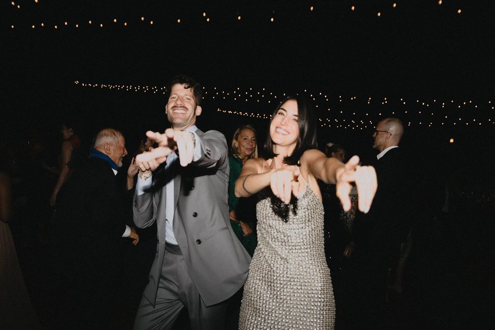 Couple dancing joyfully at a wedding reception under twinkling string lights, man in gray suit, woman in sparkly dress.