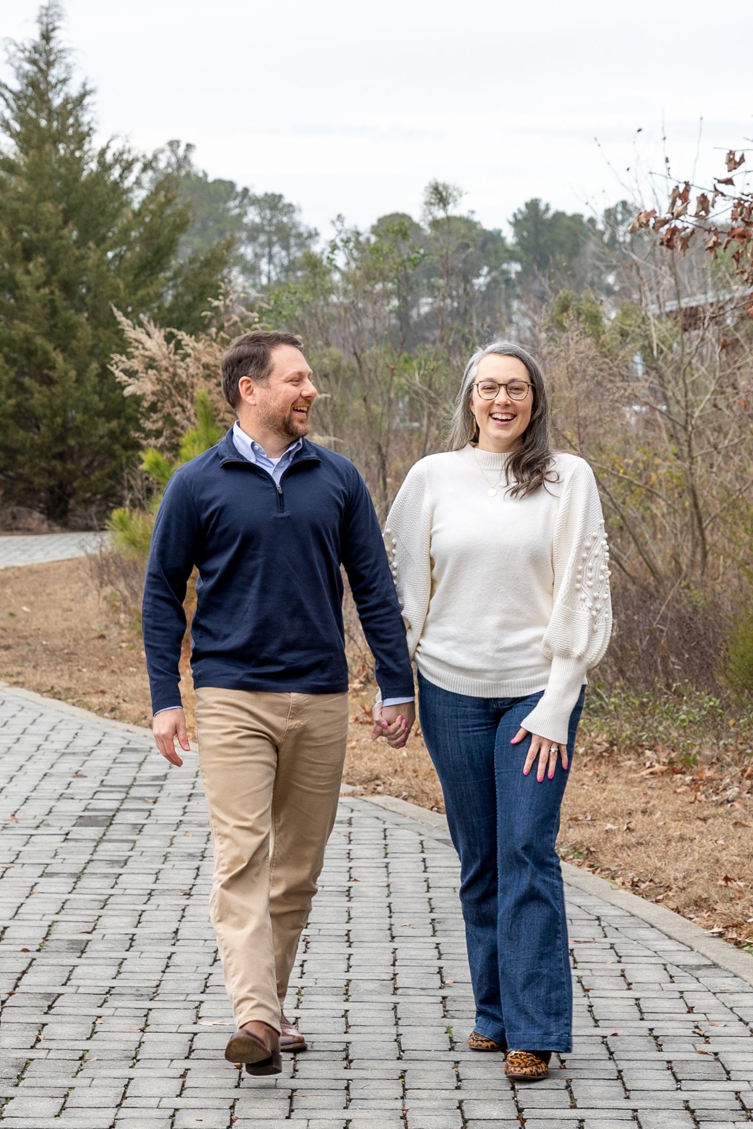 Happy couple holding hands and walking on a brick path outdoors, smiling at each other in casual winter attire.
