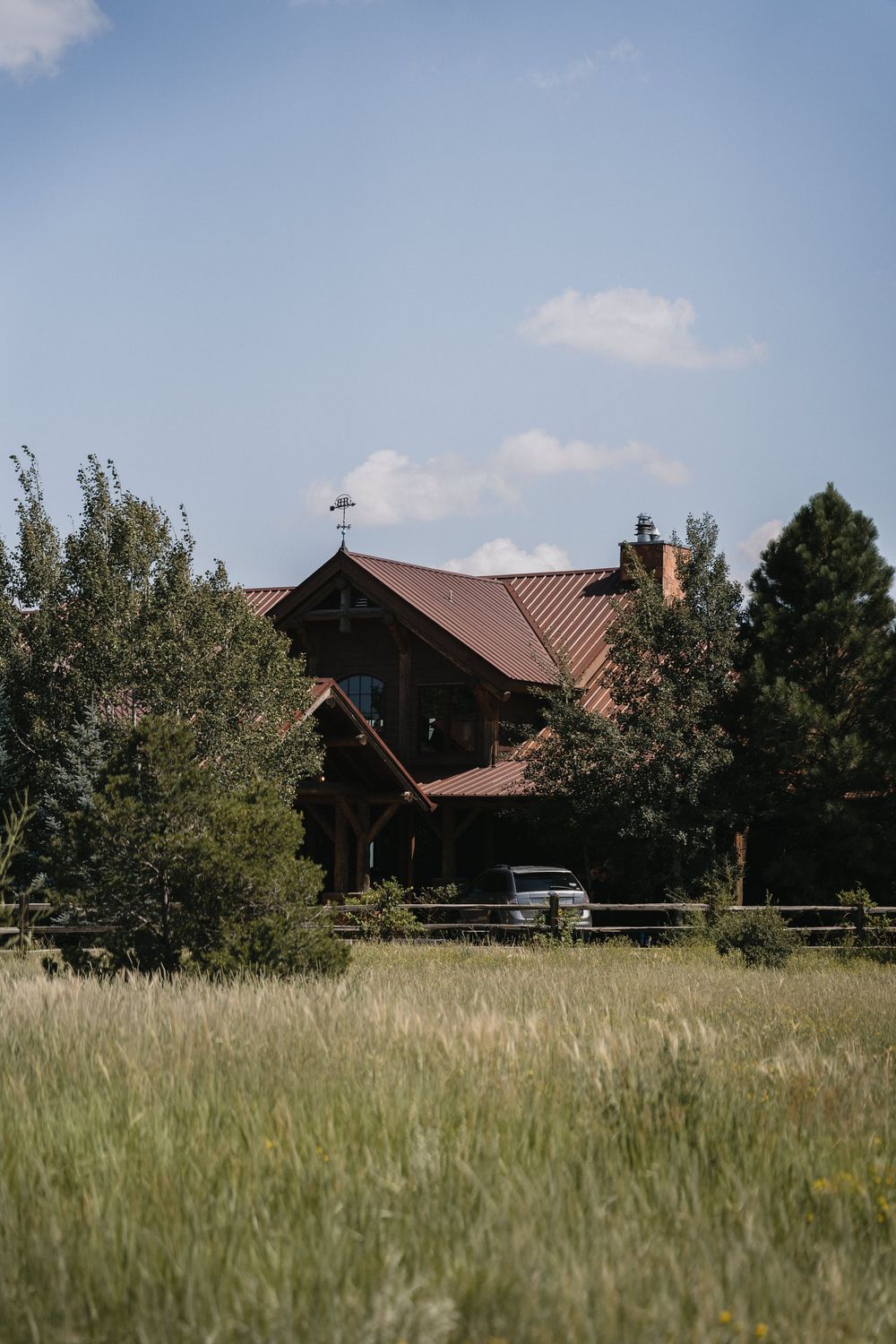 Rustic wooden lodge with red metal roof surrounded by tall trees and open green meadow under a blue sky.