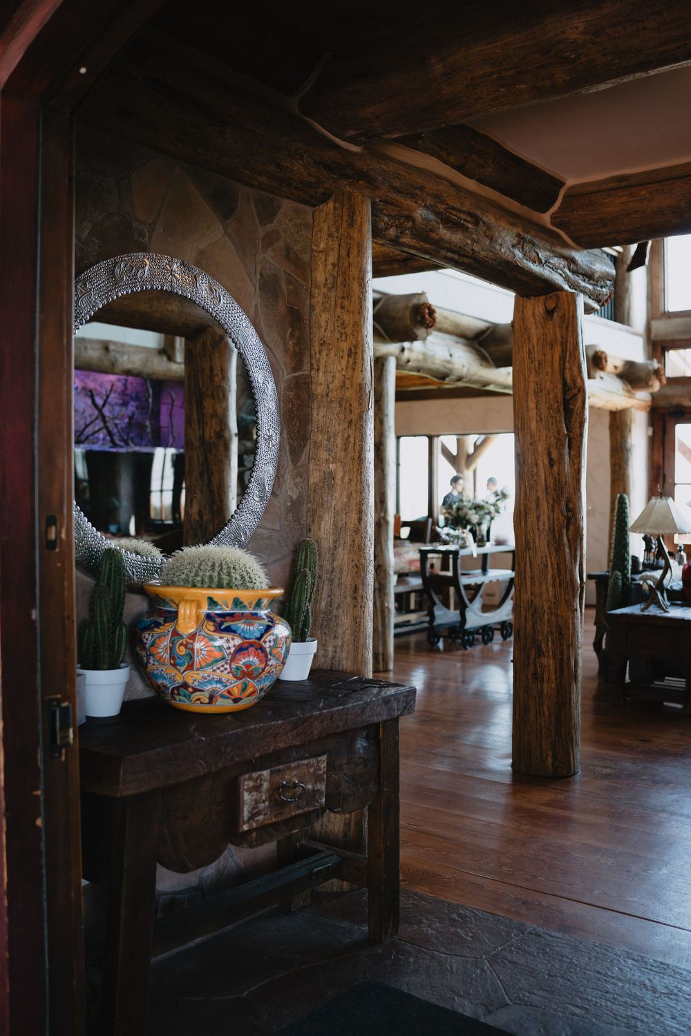 Rustic log cabin interior with colorful Talavera pottery, round carved mirror, cacti, and dark wood beams on hardwood floors.